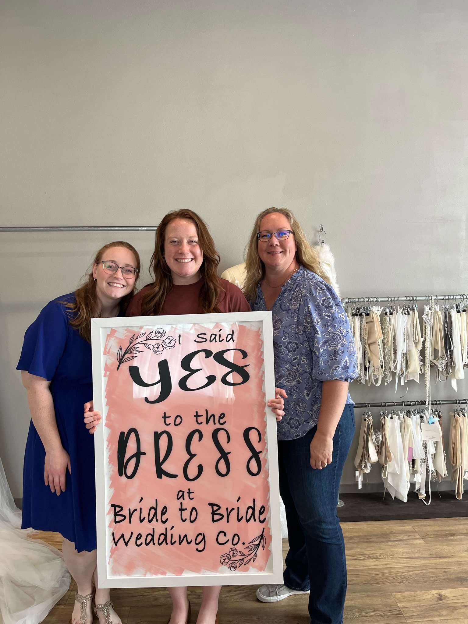 Three women at dress shop holding sign that says I said yes to the dress.
